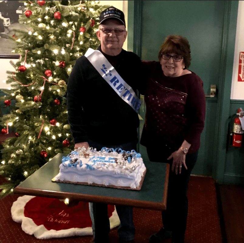 Linda and her husband David stand in front of a Christmas tree with a cake.