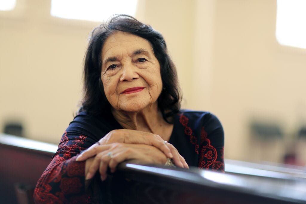 Photo of Civil Rights icon, Dolores Huerta, a woman with brown hair and brown eyes wearing a maroon and black shirt, sitting down with her hands crossed in front of her looking at the camera