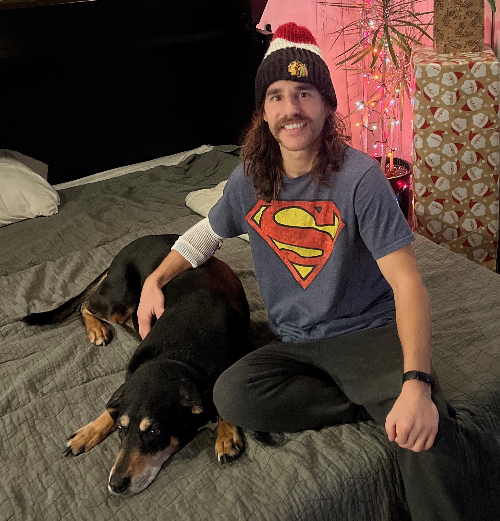 Andrew Flack, who has long wacy brown hair and a mustache wearing a superman tshirt and a red, white and blue beanie, is sitting on a bed next to his black and tan dog, Jaxson.