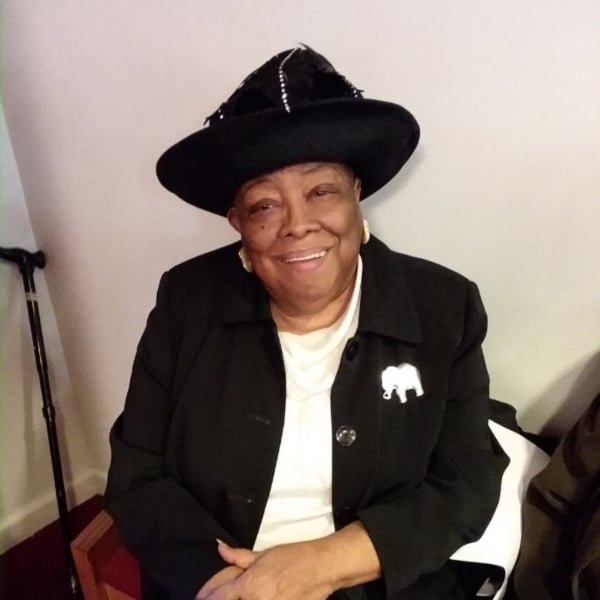 Elaine Saunders sits in a chair with her hands folded in her lap wearing a black sweater and a black hat